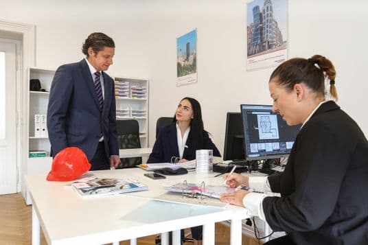 Three Engel & Völkers team members in an office discussing blueprints and a floor plan displayed on the monitor. A red hard hat, printed materials, and folders are on the desk. The scene depicts collaboration in planning or analyzing a commercial real estate project.
