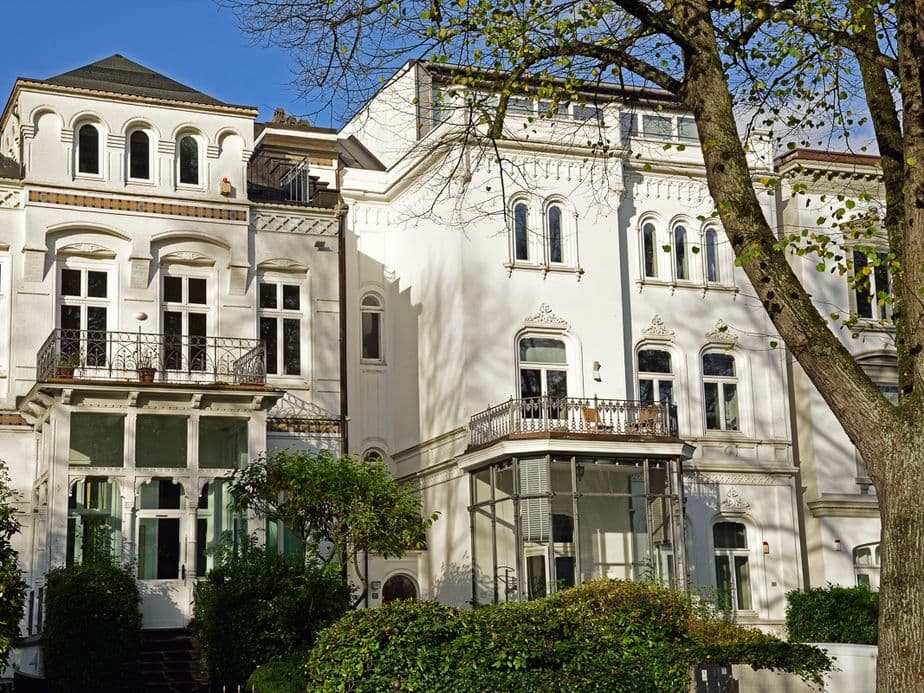 Elegant white townhouse with arched windows, balconies and a tree in the foreground under a clear blue sky.