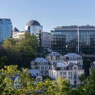 Vue urbaine de Luxembourg avec des bâtiments modernes en verre et des maisons blanches traditionnelles, encadrée par des arbres verts sous un ciel bleu.