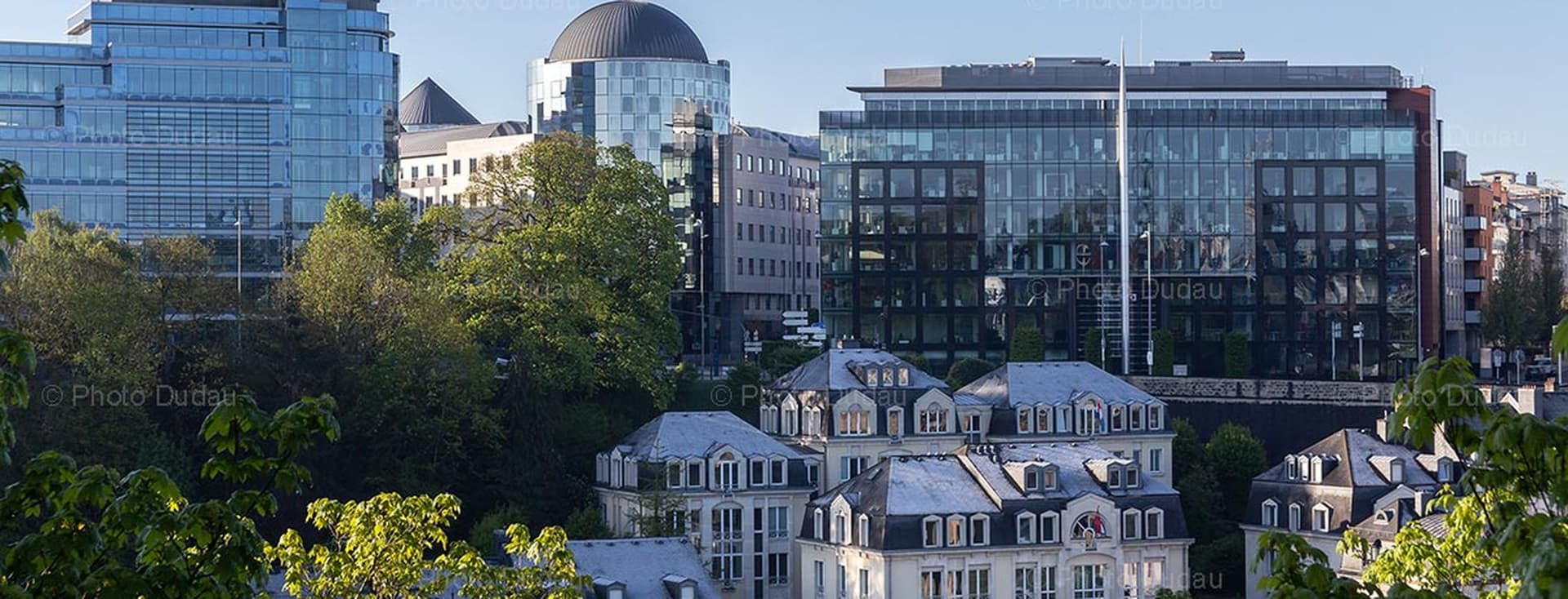 Cityscape view of Luxembourg with modern glass buildings and traditional white houses, framed by green trees under a blue sky.