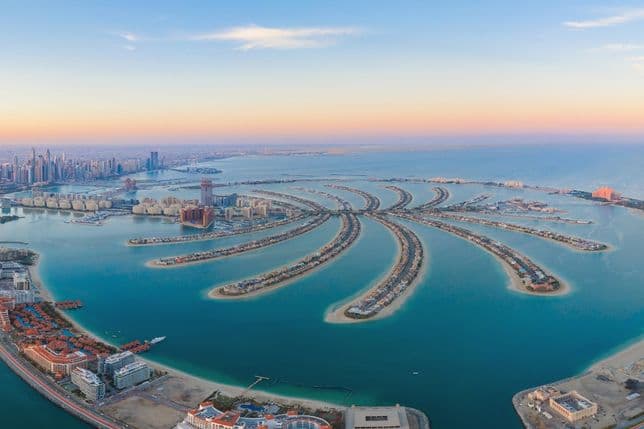 Aerial view of Palm Jumeirah in Dubai, featuring palm-shaped islands with buildings, surrounded by blue sea and distant city skyline at sunset.