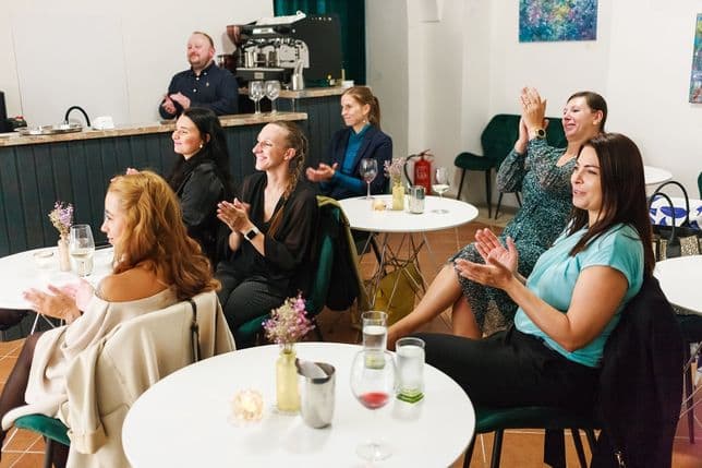 Skupina lidí sedí u kulatých stolů a tleská. V pozadí za barem stojí barista. Na stolech jsou skleničky, květiny a svíčky, které vytvářejí hřejivou a intimní atmosféru.