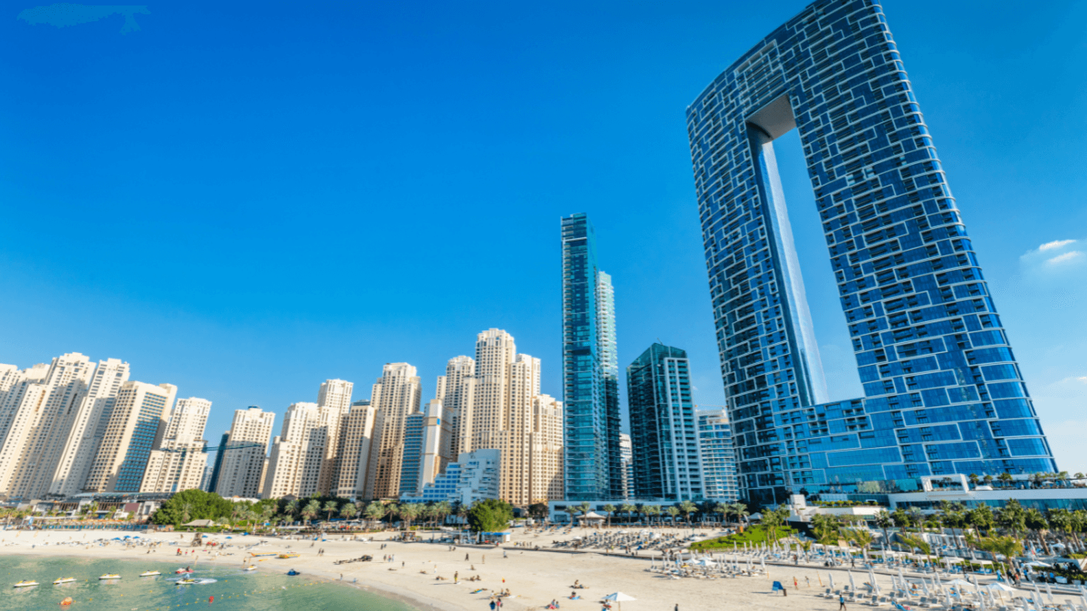 Beachfront cityscape with modern skyscrapers, including a uniquely shaped tall building, under a clear blue sky. People relax on the sandy beach.