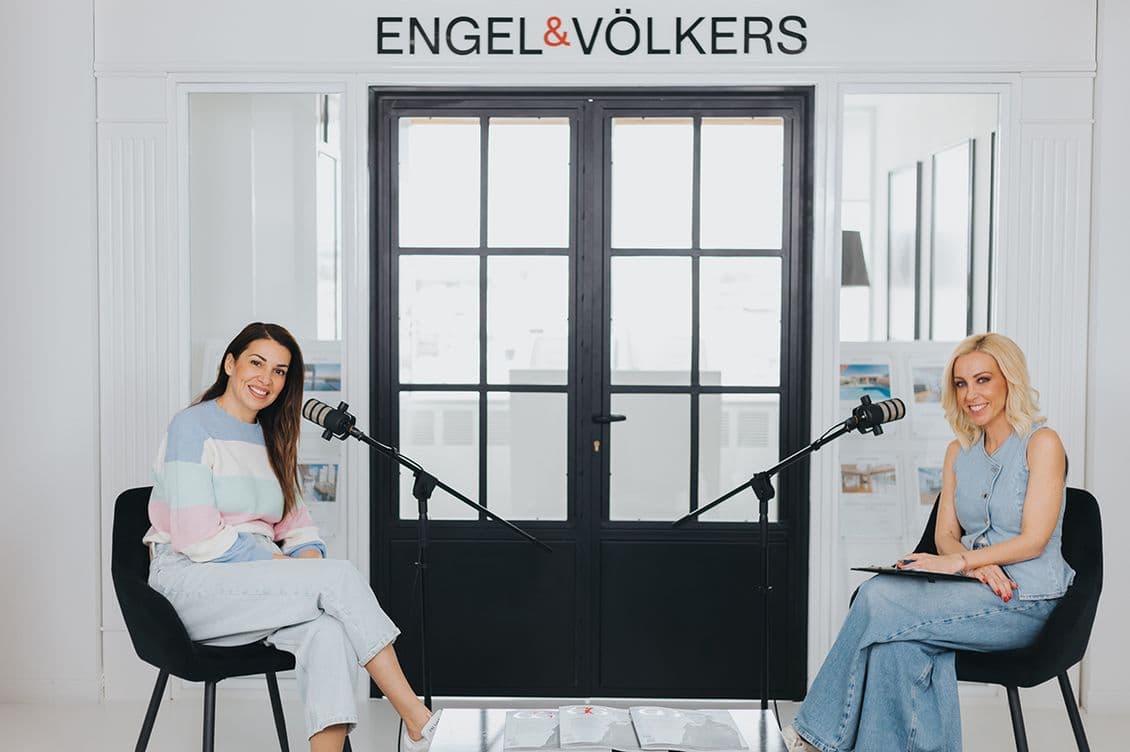 Δύο γυναίκες, η Ελένη Χατζίδου και η Λίλη Πυράκη, κάθονται σε μαύρες καρέκλες με μικρόφωνα ανάμεσά τους σε ένα φωτεινό, μοντέρνο δωμάτιο. Η πινακίδα Engel & Völkers Ελλάδος είναι ορατή πάνω από γυάλινες πόρτες με μαύρο πλαίσιο πίσω τους. Τα ποτήρια νερού στηρίζονται σε ένα μικρό τραπέζι.