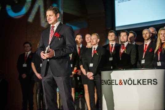 Andreas von Schlick speaks on stage in front of an audience, with the Engel & Völkers team in suits and red accents standing behind him. A professional atmosphere during a formal evening event, with the company logo visible in the foreground.
