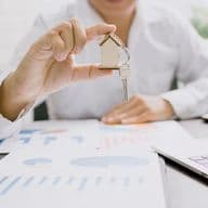 Person in a white shirt holding a small wooden house with keys attached, over a table with graphs and charts. A laptop is partially visible. The setting suggests a business or real estate context.