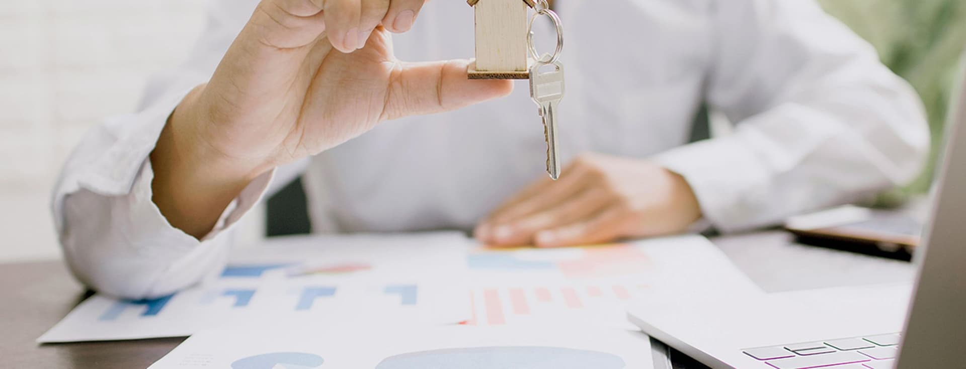 Person in a white shirt holding a small wooden house with keys attached, over a table with graphs and charts. A laptop is partially visible. The setting suggests a business or real estate context.