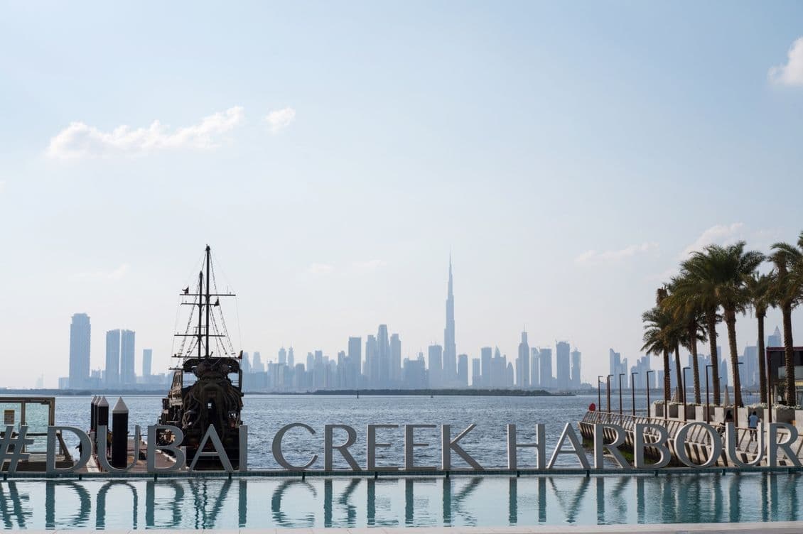 Skyline of Dubai with Burj Khalifa in the distance, seen from Dubai Creek Harbour. A traditional boat and palm trees are in the foreground.