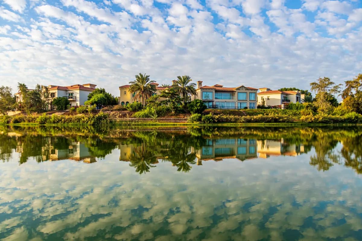 Lakeside view of modern houses with palm trees, reflected in calm water under a sky filled with scattered clouds.