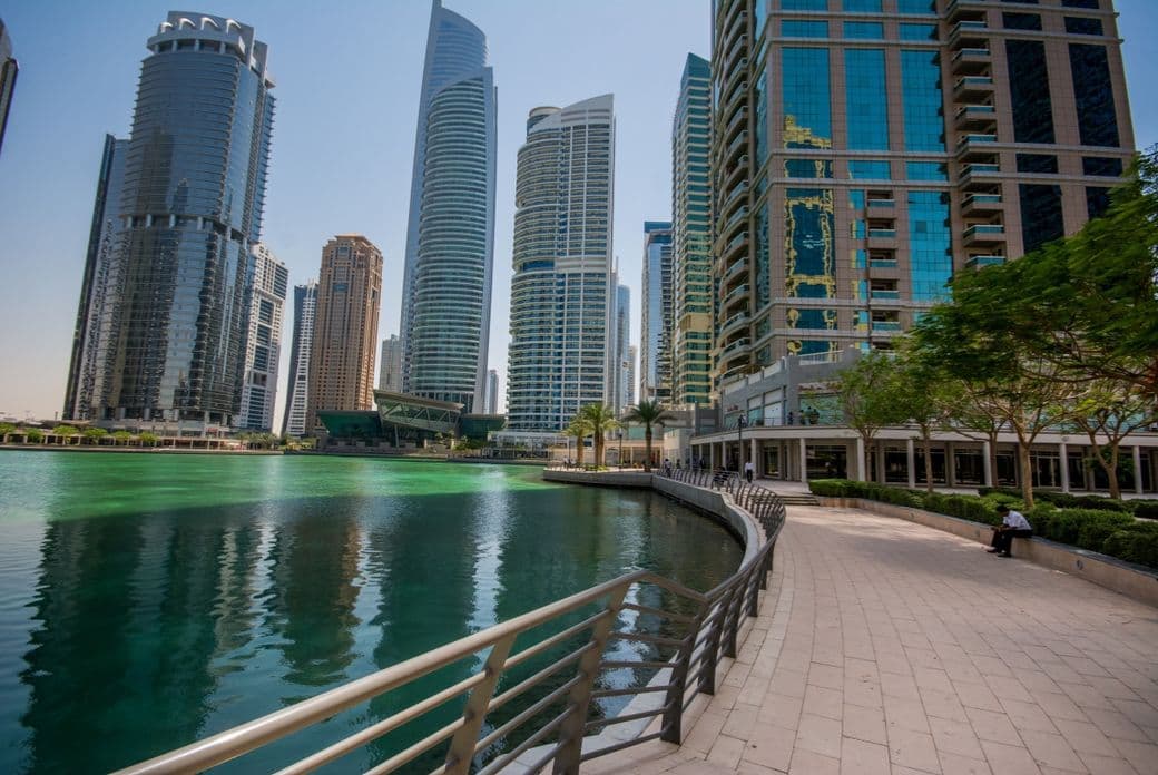 A curved walkway beside a turquoise lake, surrounded by modern skyscrapers and lush trees under a clear blue sky.
