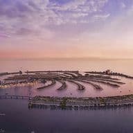 Aerial view of Palm Jebel Ali at sunset, surrounded by calm sea, with distant cityscape and a soft, colorful sky.