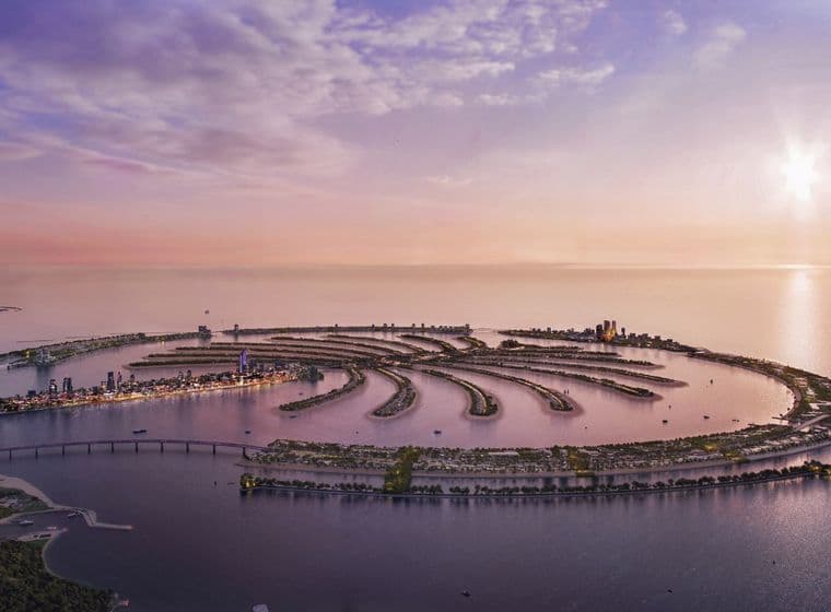 Aerial view of Palm Jebel Ali at sunset, surrounded by calm sea, with distant cityscape and a soft, colorful sky.