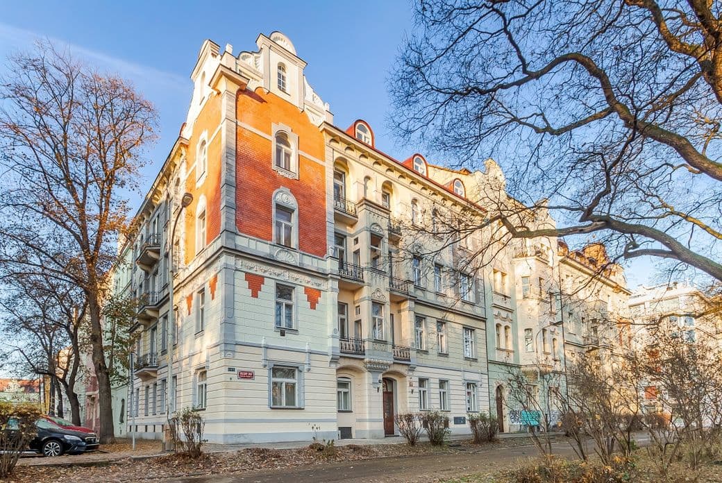 The photo shows a four-storey historic apartment building situated on a corner plot. The façade combines light cream tones with brick-red accents, and features balconies, bay windows, and decorative elements typical of early 20th-century architecture. Leafless trees frame the scene, with a walkway and a few parked cars visible in the foreground.