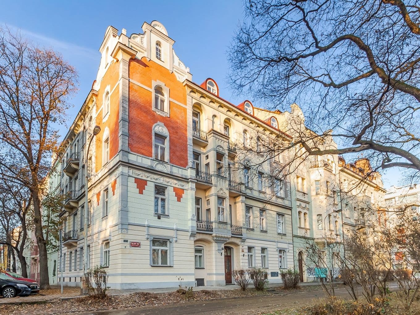 The photo shows a four-storey historic apartment building situated on a corner plot. The façade combines light cream tones with brick-red accents, and features balconies, bay windows, and decorative elements typical of early 20th-century architecture. Leafless trees frame the scene, with a walkway and a few parked cars visible in the foreground.