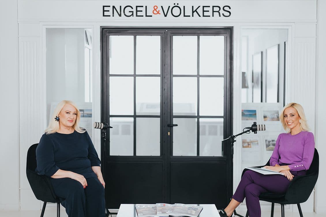 Two women seated in a modern office with "Engel & Völkers" sign above. They are smiling, with microphones and magazines on a table between them.
