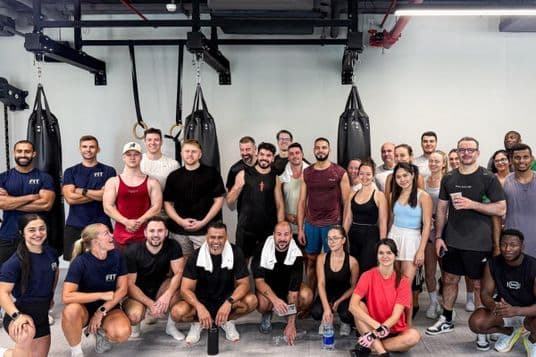A diverse group of people smiling and posing in a gym, surrounded by punching bags and fitness equipment.