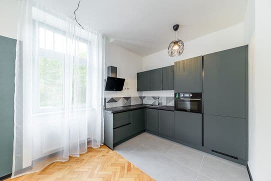 Kuchyňský kout s tmavě zelenými skříňkami, vestavěnou troubou, varnou deskou a digestoří, doplněný velkým oknem se záclonami.