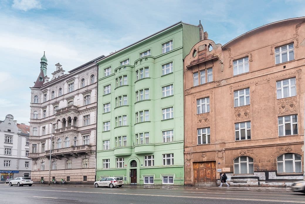 View of a row of historic city buildings along a wide street. In the center stands a multi-storey apartment building with a light green façade and rounded bay windows. On both sides are ornate buildings in light and sand colors with decorative details and tall windows. In front of the buildings runs a road with passing cars and a sidewalk where a pedestrian is walking.