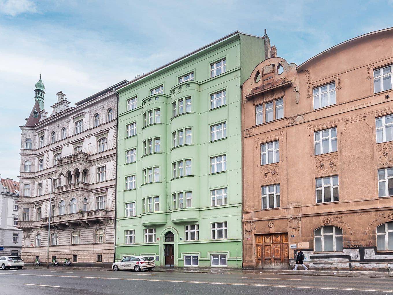 View of a row of historic city buildings along a wide street. In the center stands a multi-storey apartment building with a light green façade and rounded bay windows. On both sides are ornate buildings in light and sand colors with decorative details and tall windows. In front of the buildings runs a road with passing cars and a sidewalk where a pedestrian is walking.