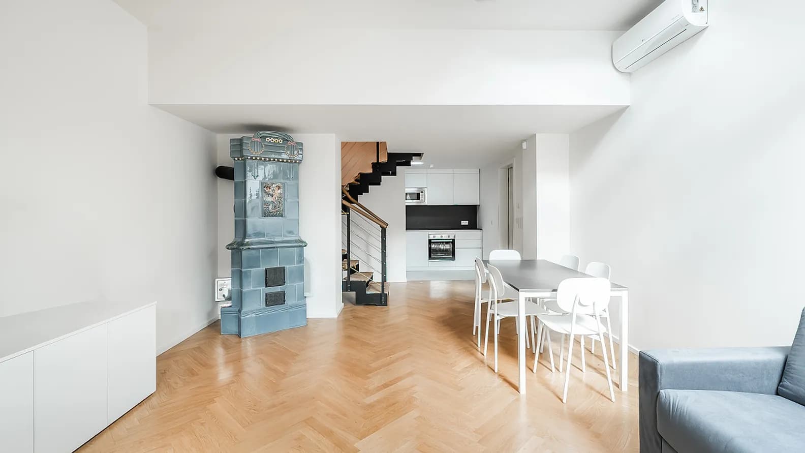 Bright living space with parquet flooring, a dining area, kitchen, staircase and original tiled stove.