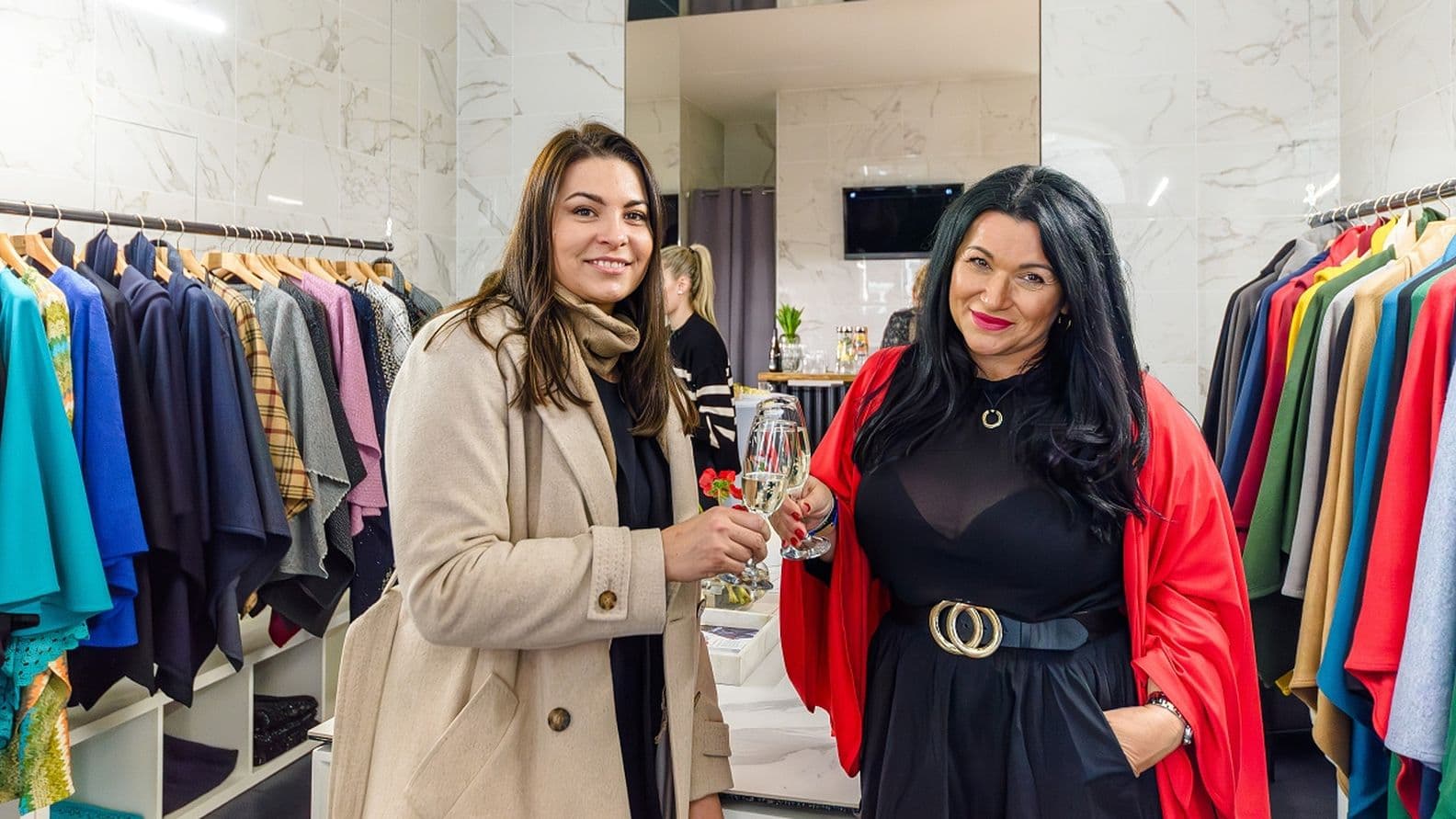 Dvě ženy si připíjejí sklenkou šampaňského ve stylovém módním butiku, v pozadí jsou stojany s barevným dámským oblečením a zrcadlová stěna.