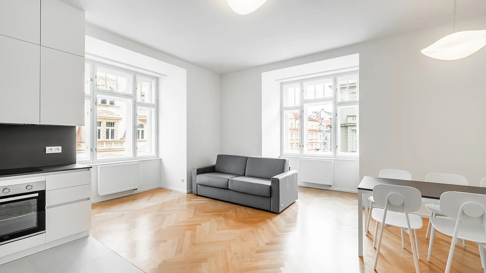 Bright living room with a modern kitchen, dining area, grey sofa, parquet flooring and large windows.