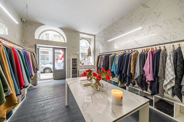 Interiér stylového butiku se dvěma řadami barevných dámských kabátů, mramorovými stěnami, zapálenou svíčkou a květinami na centrálním stole. Otevřené dveře vedou ven na ulici.