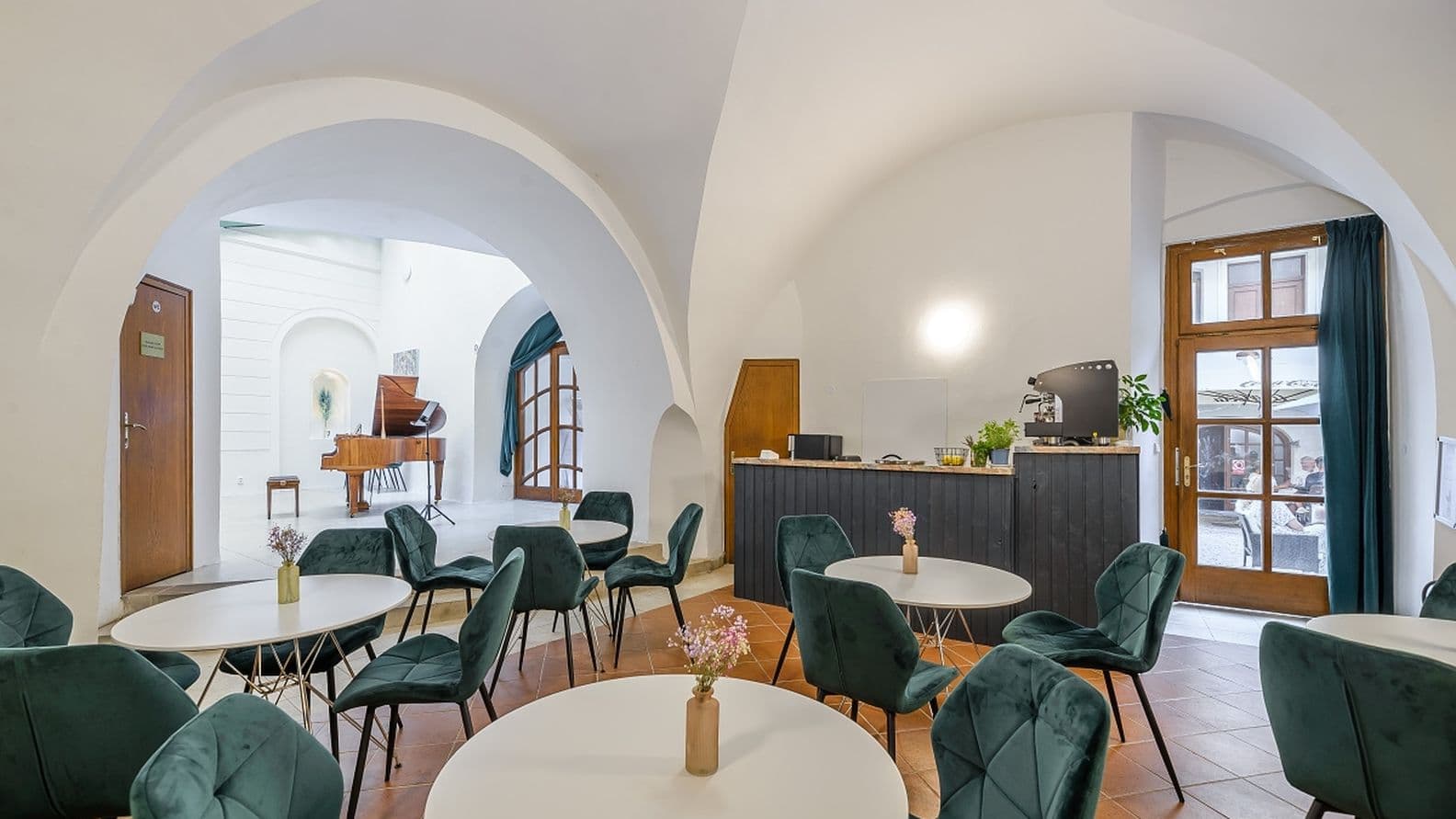 Interiér galerie s klenutými stropy a elegantním posezením. V pozadí je vidět koncertní křídlo, v popředí kulaté stolky s vázami květin.