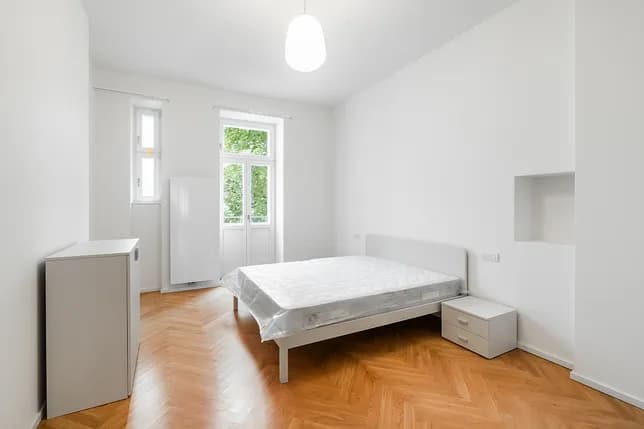 Bright bedroom with a double bed, white furniture, wooden parquet flooring and tall windows overlooking greenery.