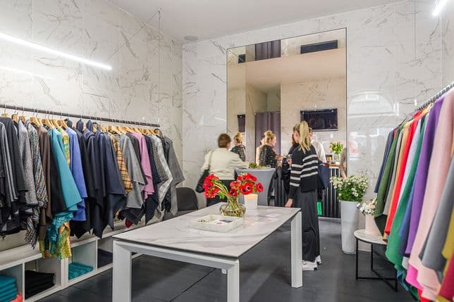 Elegantní butik s mramorovými stěnami, centrálním stolem s květinami a barevným dámským oblečením na stojanech. V pozadí si u pultu povídá několik žen.
