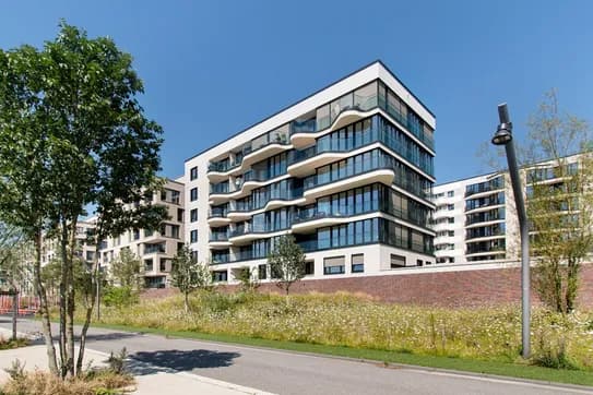 Modernes Wohnhaus mit geschwungenen Glasbalkonen und einer weißen Fassade, mit Blick auf eine Grünfläche mit Bäumen und einer Backsteinmauer unter blauem Himmel.