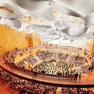 Interior view of a concert hall with wood walls, white ceiling panels, and tiered seating filled with people.