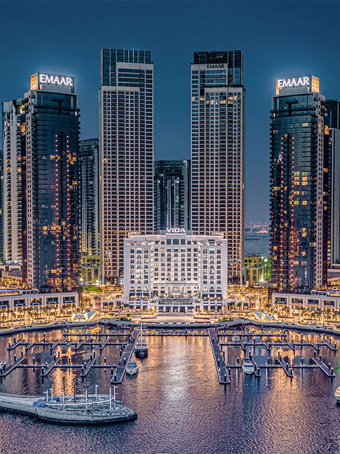 Night view of illuminated skyscrapers and marina with boats docked, reflecting on calm water. Buildings display "EMAAR" signs at the top.