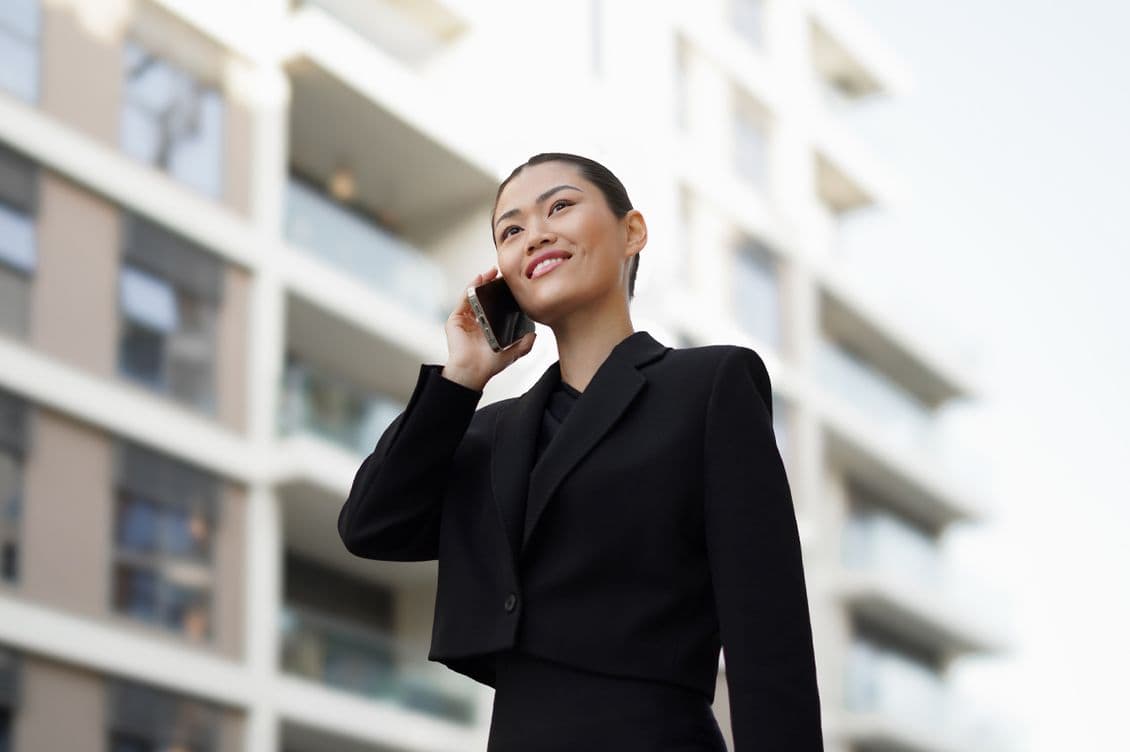 Woman in a black suit smiling while talking on a phone, standing in front of a modern apartment building.