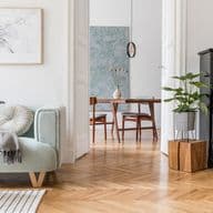 Cozy living area featuring a mint-green accent chair with throw blanket, abstract wall art, vintage black piano, herringbone wooden flooring, and modern dining area visible through doorway, blending contemporary and traditional elements