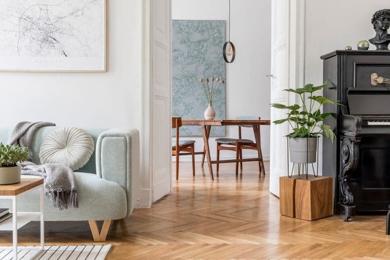 Cozy living area featuring a mint-green accent chair with throw blanket, abstract wall art, vintage black piano, herringbone wooden flooring, and modern dining area visible through doorway, blending contemporary and traditional elements