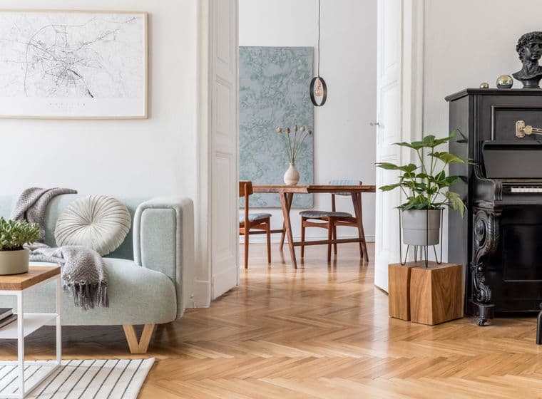 Cozy living area featuring a mint-green accent chair with throw blanket, abstract wall art, vintage black piano, herringbone wooden flooring, and modern dining area visible through doorway, blending contemporary and traditional elements