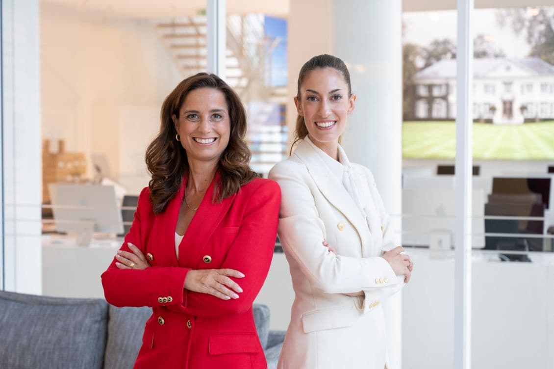 Two women smiling confidently, standing with arms crossed in a modern office space. One wears a red suit, the other a white suit.