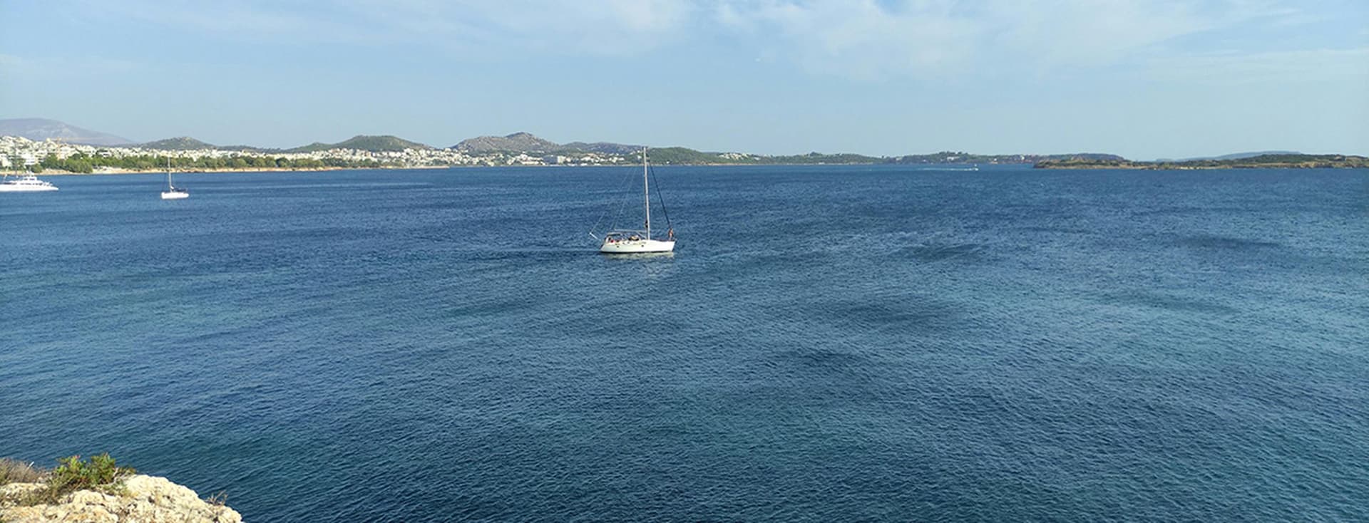 A sailboat on a calm blue sea near a rocky shore, with distant hills and a town visible under a partly cloudy sky.