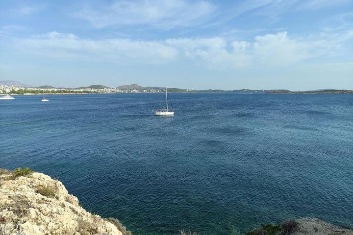A sailboat on a calm blue sea near a rocky shore, with distant hills and a town visible under a partly cloudy sky.