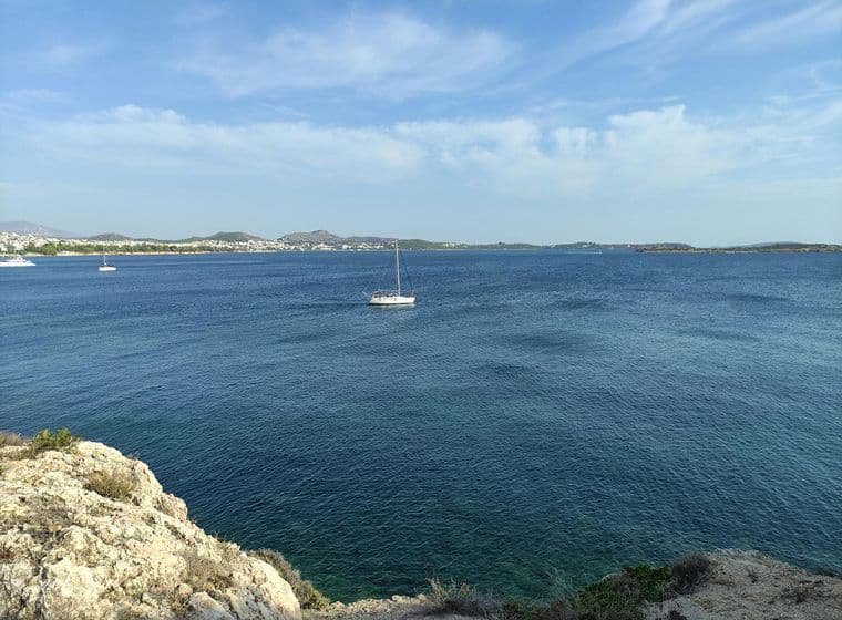 A sailboat on a calm blue sea near a rocky shore, with distant hills and a town visible under a partly cloudy sky.