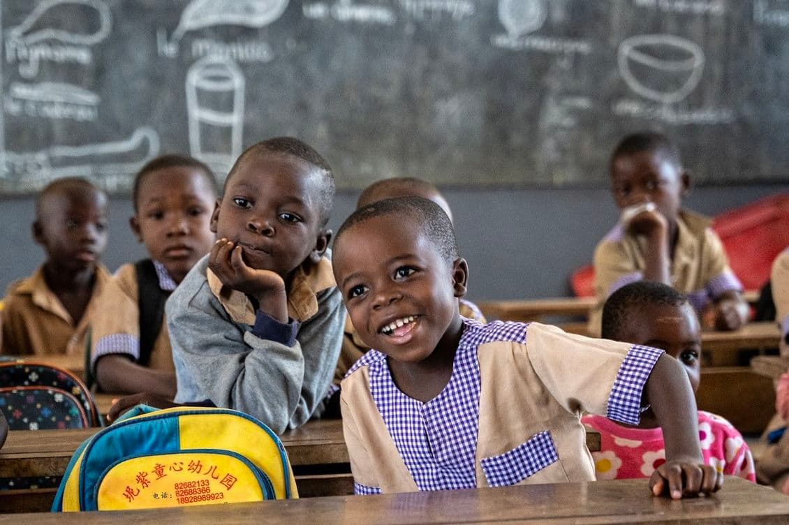 Smiling children in a classroom focus on their lessons while sitting at wooden desks, their backpacks beside them. A chalkboard with writing is visible in the background, capturing a lively learning environment.
