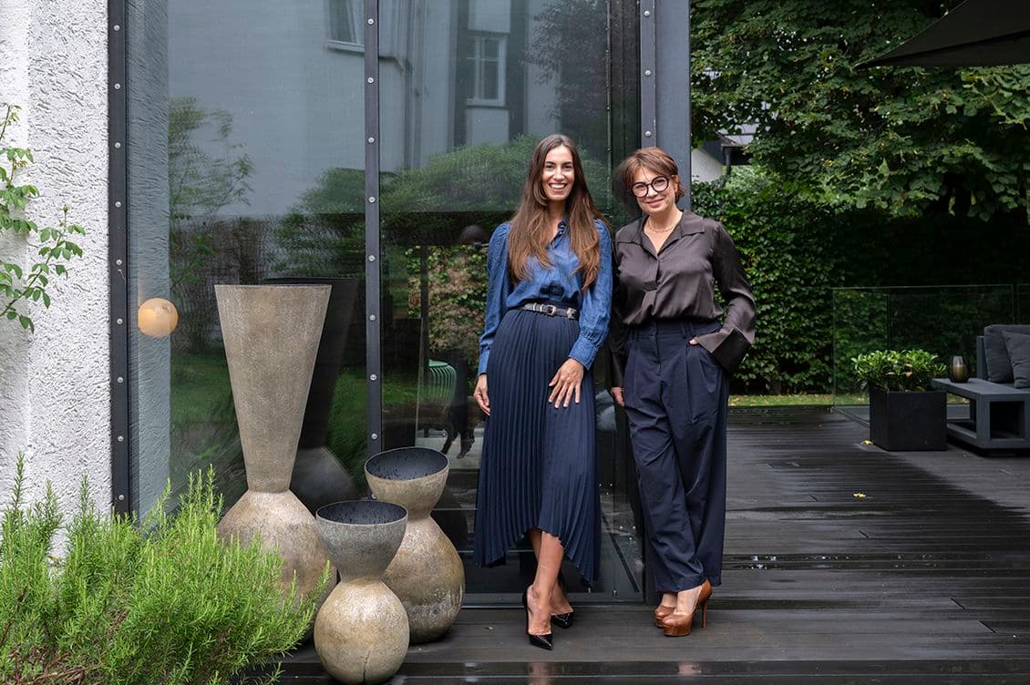 Two women stand on a wooden deck beside large vases and greenery, with a modern glass building in the background.