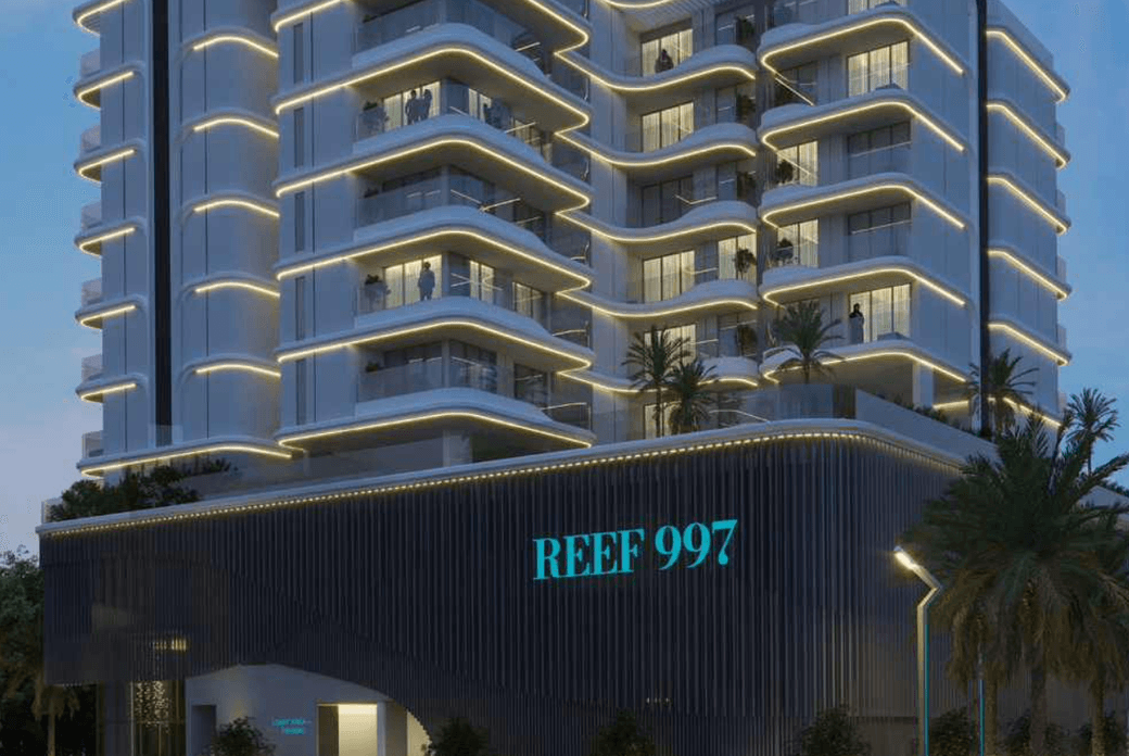 Modern multi-story building labeled "REEF 997" with illuminated balconies and palm trees, set against an evening sky.