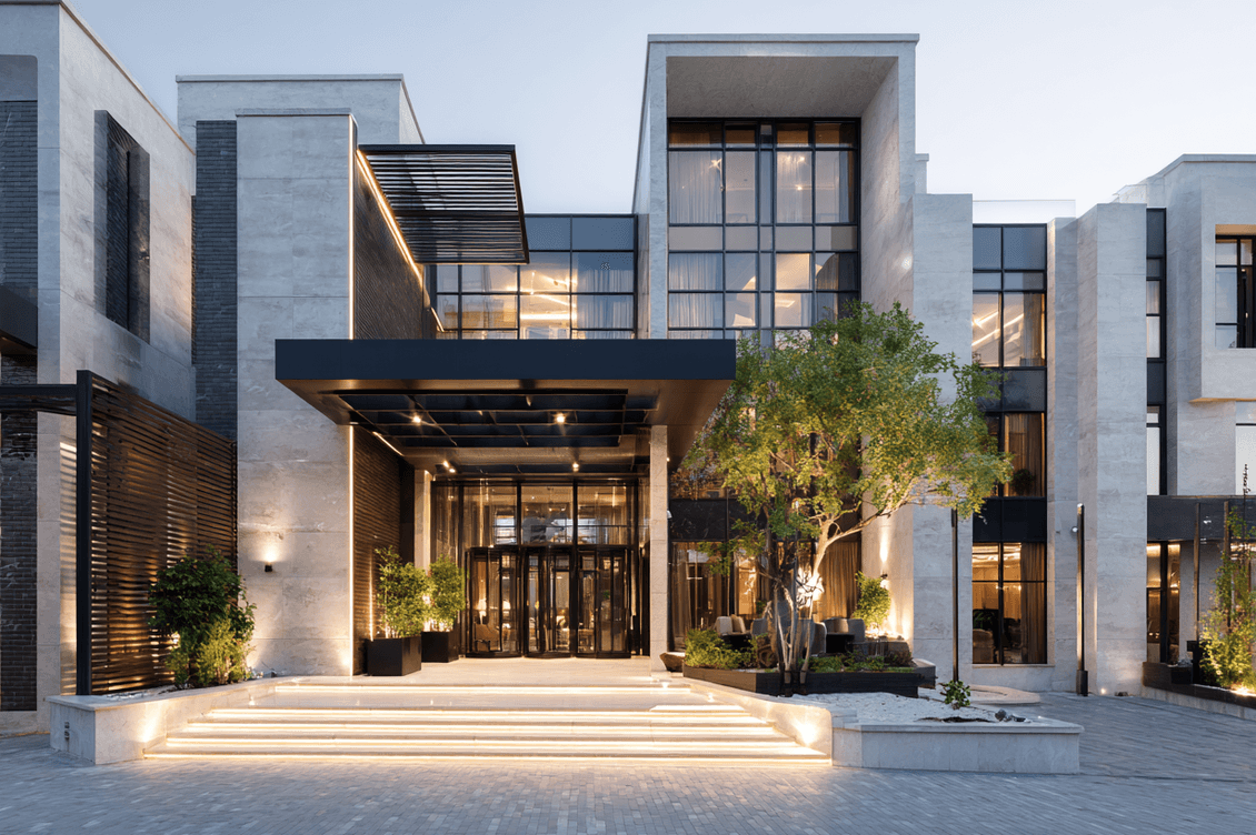 Modern building with large glass windows, illuminated entrance, greenery, and steps leading up to a spacious canopy. Evening lighting enhances design.