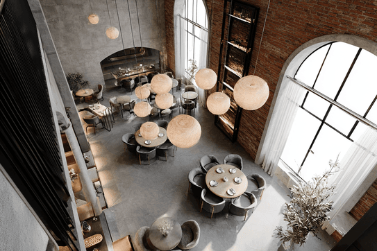 Aerial view of a modern restaurant with round tables, plush chairs, large arched windows, hanging globe lights, and exposed brick walls.