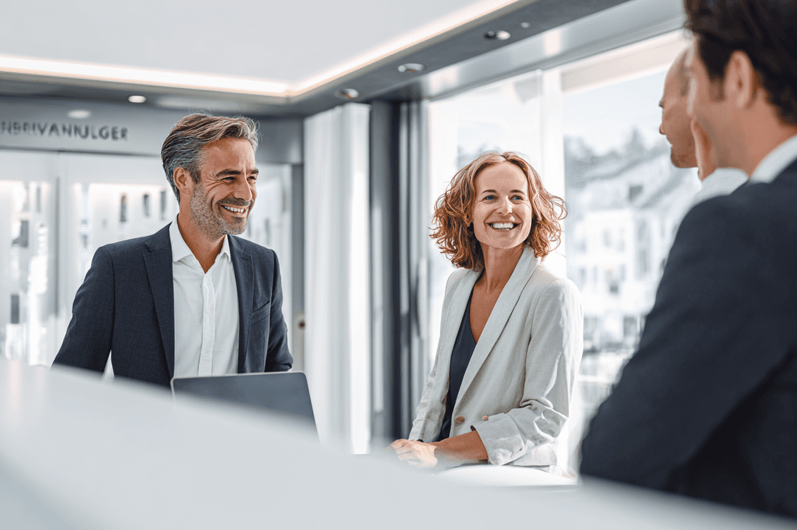 A group of professionals in a modern office, engaging in a friendly discussion with smiles, against a backdrop of large windows.