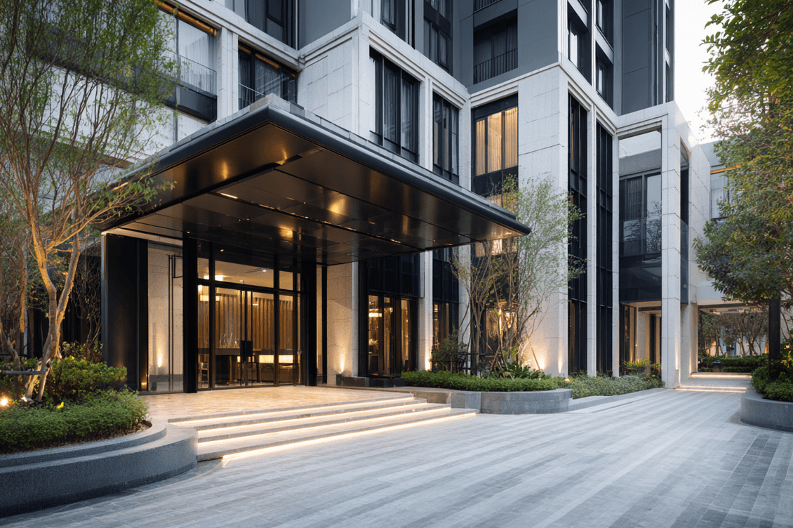 Modern building entrance with large glass doors, landscaped walkway, and ambient lighting. Surrounded by trees and low shrubs.