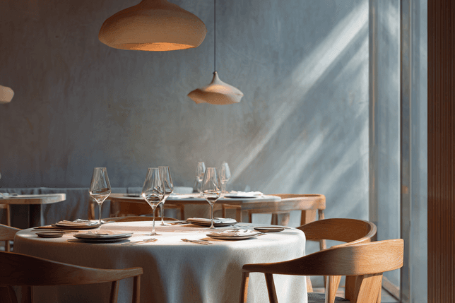 Elegant restaurant interior with round tables, white tablecloths, wooden chairs, and pendant lighting, softly lit by natural light from large windows.