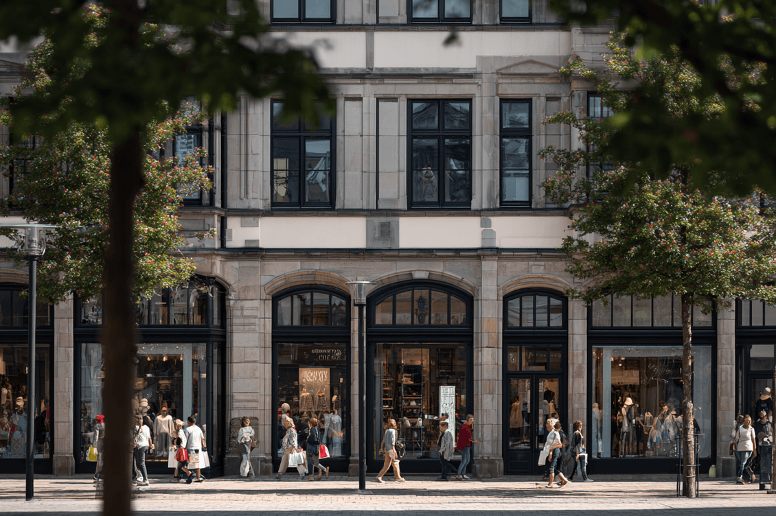 A group of people walk past a stylish building with large windows and arched doorways. Trees frame the scene in an urban setting.